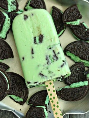 A White Chocolate Mint Cookie Popsicle laying on a pan full of broken Mint Oreo Cookies.
