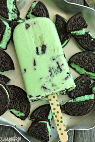 A White Chocolate Mint Cookie Popsicle laying on a pan full of broken Mint Oreo Cookies.