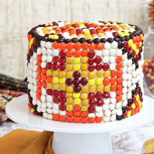 Patterned M&M Cake on a white cake plate next to an orange napkin and dried corn.