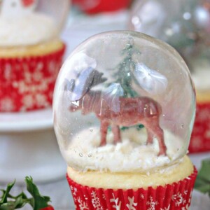 A snow globe cupcake with a moose and a tree on it and other snow globe cupcakes in the background.
