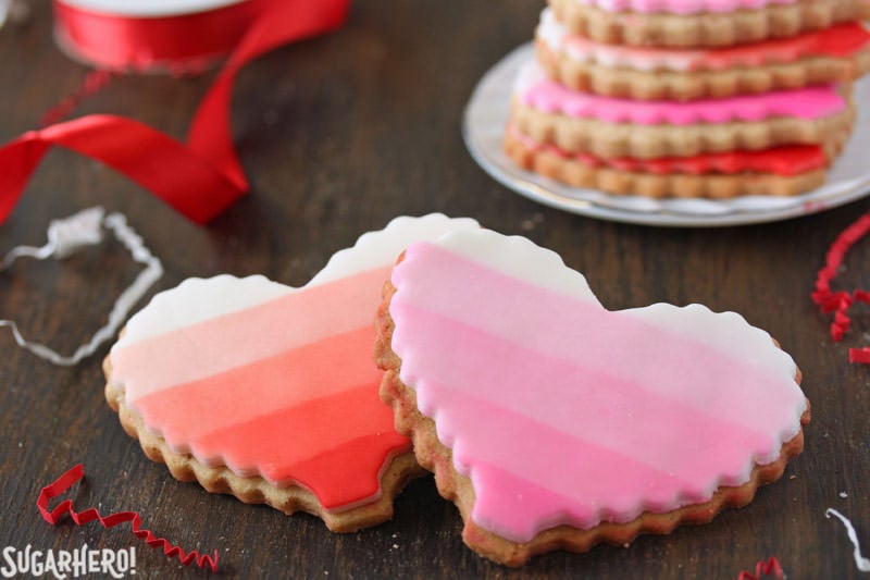Brown Butter Heart Cookies - brown butter sugar cookies with pretty patterns for Valentine's Day! | From SugarHero.com Brown Butter Heart Cookies - two sugar cookies, one red ombre and one pink ombre, with a stack of cookies behind | From SugarHero.com