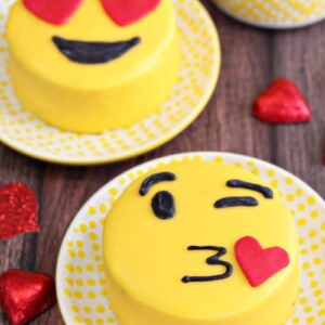Two Emoji Cakes, one with a kissy face and one with heart eyes, on yellow polka dotted plates.