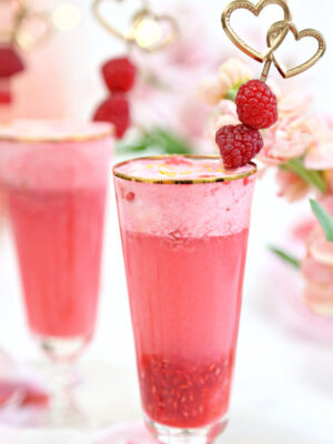 Two gold-rimmed glasses with pink raspberry punch and a gold raspberry swizzle stick garnish.