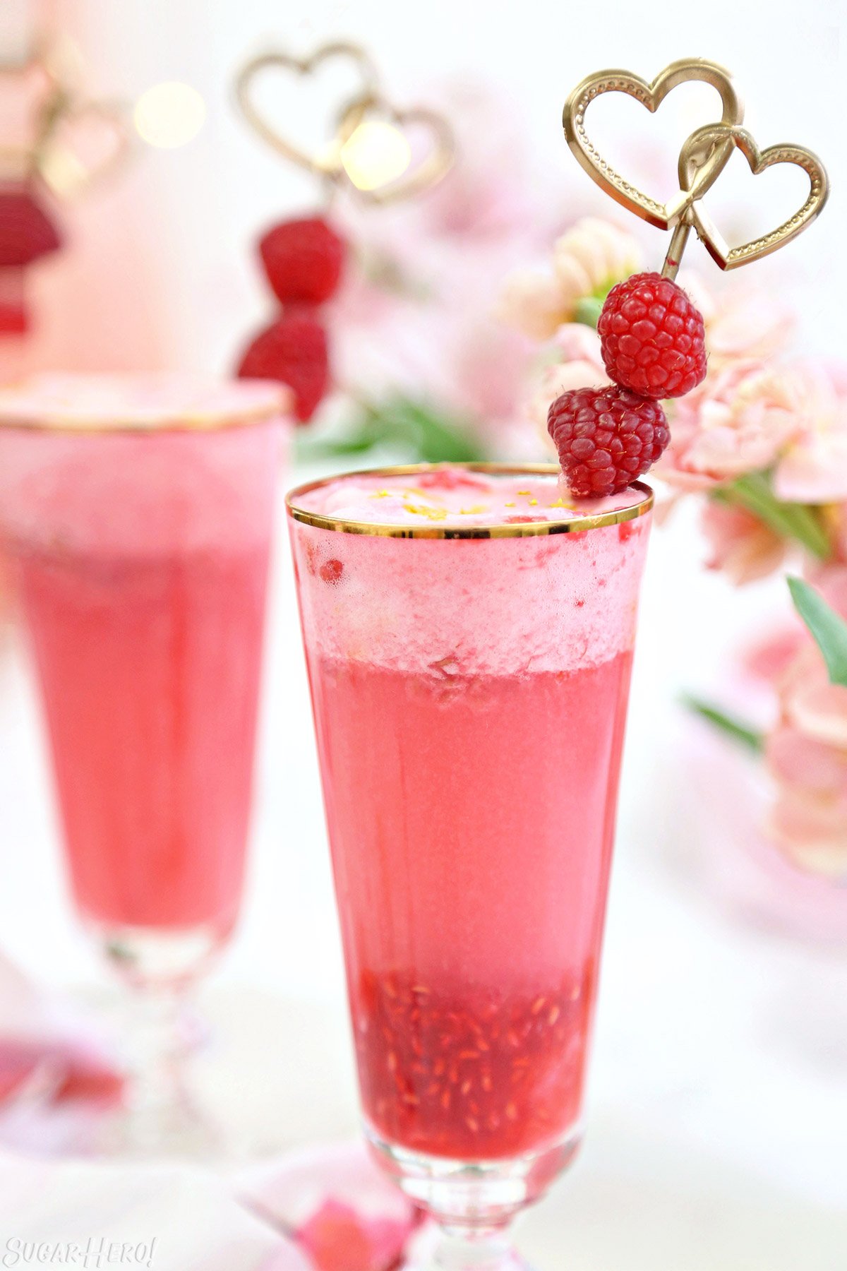 Two gold-rimmed glasses with pink raspberry punch and a gold raspberry swizzle stick garnish.