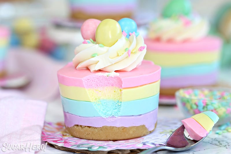Easter No-Bake Mini Cheesecakes - pastel striped cheesecakes that are super easy, no baking required! | From SugarHero.com Easter no-bake mini cheesecake topped with frosting, sprinkles, and a candy egg. There is a a spoonful missing and set beside the cheesecake.