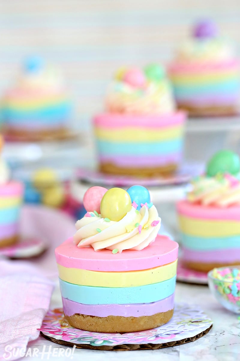 Easter no-bake mini cheesecakes topped with frosting, sprinkles, and candy eggs on colorful doilies.