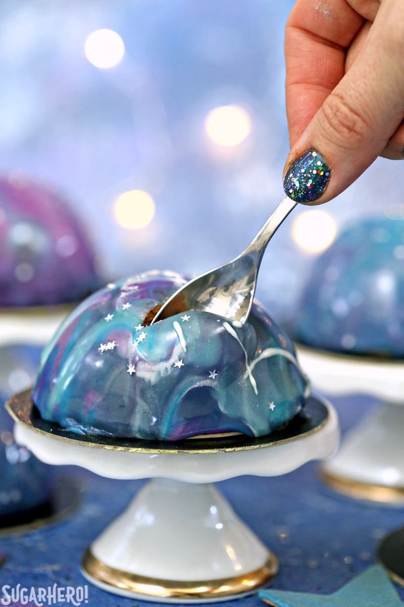 Galaxy Mousse Cakes are almost too pretty to be real! Mini chocolate mousse cakes on a brownie base are covered with a gorgeous glossy mirror glaze that’s swirled to look like the galaxy. They are delicious, edible works of art! | From SugarHero.com Galaxy Mousse Cakes - A close up shot of a mousse cake with a spoon scooping out a bite. | From SugarHero.com