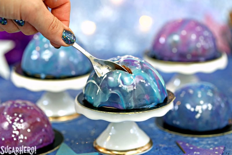 Galaxy Mousse Cakes are almost too pretty to be real! Mini chocolate mousse cakes on a brownie base are covered with a gorgeous glossy mirror glaze that’s swirled to look like the galaxy. They are delicious, edible works of art! | From SugarHero.com Galaxy Mousse Cakes - A close up shot of a mousse cake with a spoon digging in. | From SugarHero.com