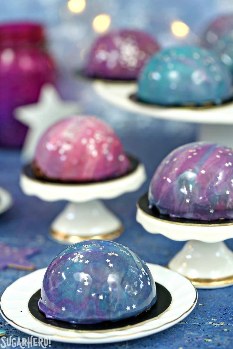 Galaxy Mousse Cakes are almost too pretty to be real! Mini chocolate mousse cakes on a brownie base are covered with a gorgeous glossy mirror glaze that’s swirled to look like the galaxy. They are delicious, edible works of art! | From SugarHero.com Galaxy Mousse Cakes - Mini Mousse Galaxy Cakes displayed on mini cake stands. | From SugarHero.com