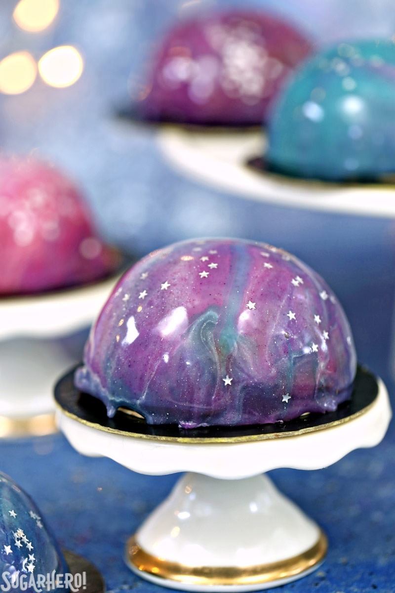 Galaxy Mousse Cakes are almost too pretty to be real! Mini chocolate mousse cakes on a brownie base are covered with a gorgeous glossy mirror glaze that’s swirled to look like the galaxy. They are delicious, edible works of art! | From SugarHero.com Galaxy Mousse Cakes - A close up shot of a mini mousse cake. | From SugarHero.com