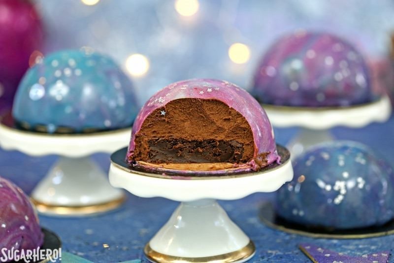 Galaxy Mousse Cakes are almost too pretty to be real! Mini chocolate mousse cakes on a brownie base are covered with a gorgeous glossy mirror glaze that’s swirled to look like the galaxy. They are delicious, edible works of art! | From SugarHero.com Galaxy Mousse Cakes - A mini mousse cake with a bike taken out, showing the inside. | From SugarHero.com