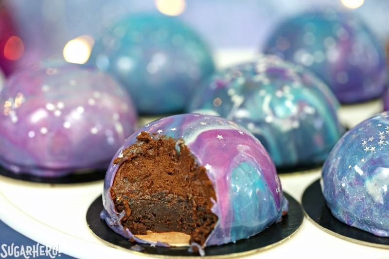 Galaxy Mousse Cakes are almost too pretty to be real! Mini chocolate mousse cakes on a brownie base are covered with a gorgeous glossy mirror glaze that’s swirled to look like the galaxy. They are delicious, edible works of art! | From SugarHero.com Galaxy Mousse Cakes - A close up shot of a mousse cake with a bite taken out of it. | From SugarHero.com
