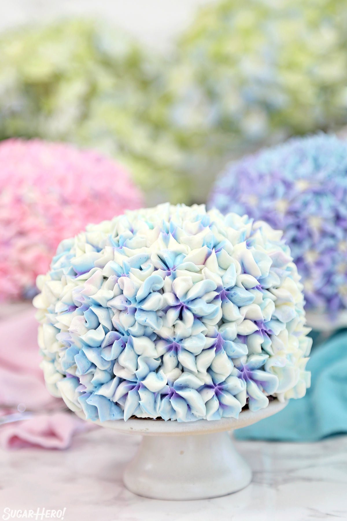 Hydrangea Cakes - A close up shot of a cake on a cake stand with blurred ones in the back. | From SugarHero.com