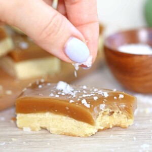 Hand sprinkling salt on a Salted Caramel Bar with a bite taken out of it.