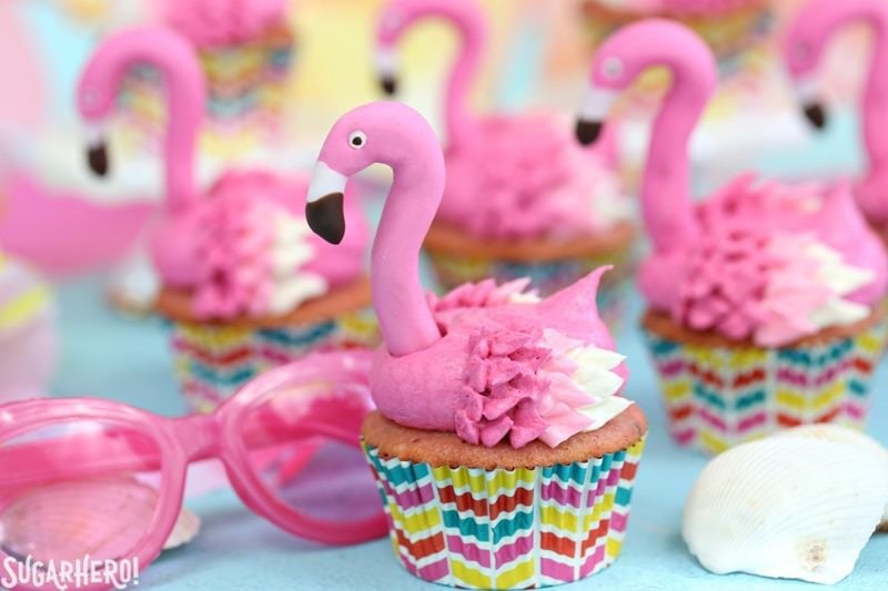 Flamingo Cupcakes - pink lemonade cupcakes decorated to look like pink flamingos! Easy, cute, and perfect for summer! | From SugarHero.com Group of Flamingo Cupcakes next to pink sunglasses and a seashell