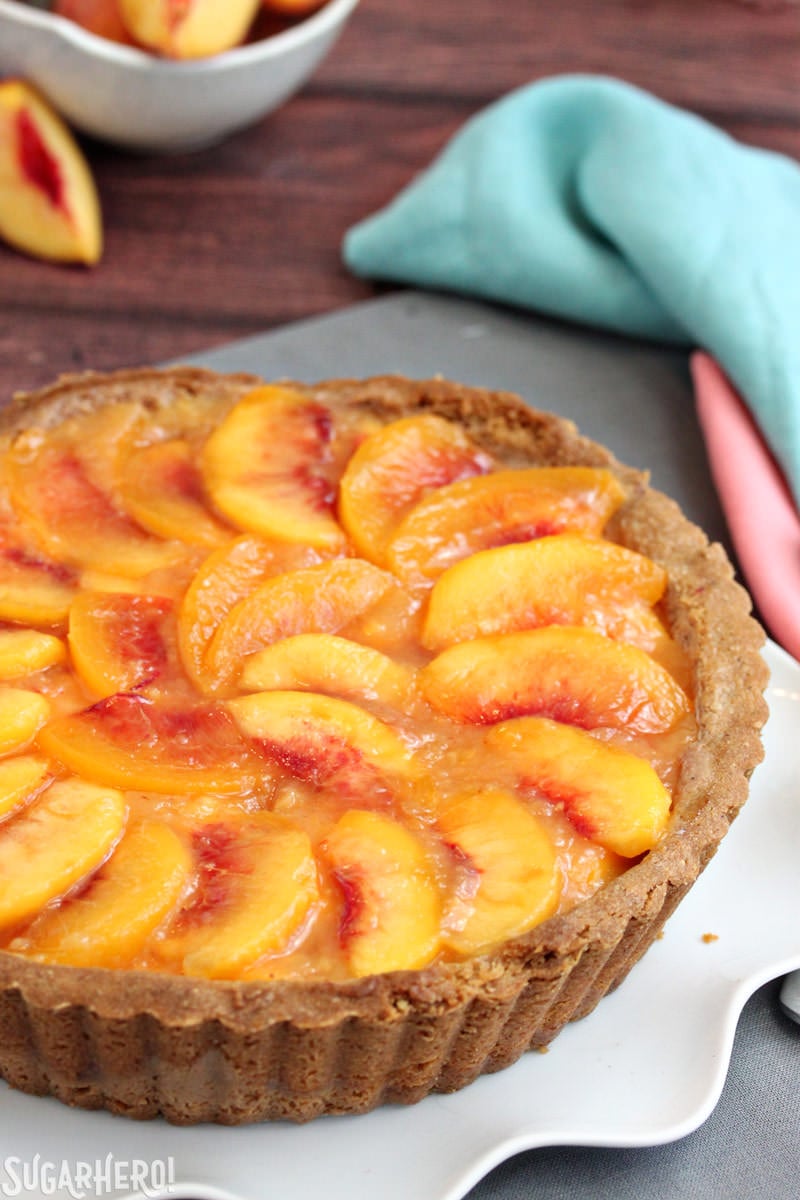 Fresh Peach Tart - the BEST way to enjoy peaches! No-bake tart recipe featuring juicy ripe peaches in a buttery shell | From SugarHero.com Vertical image of a no-bake Fresh Peach Tart with peaches in the background | From SugarHero.com