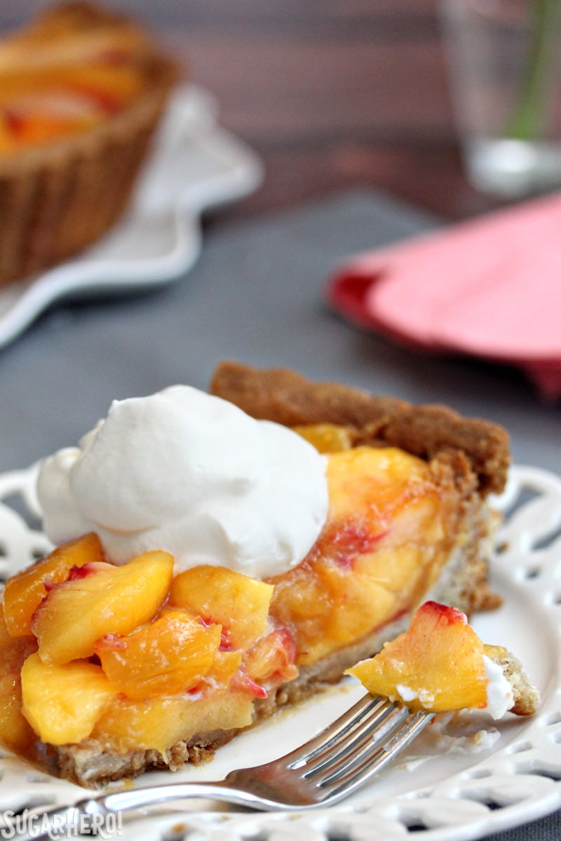 Fresh Peach Tart - the BEST way to enjoy peaches! No-bake tart recipe featuring juicy ripe peaches in a buttery shell | From SugarHero.com A sslice of no-bake Fresh Peach Tart, topped with whipped cream, with the whole tart in the background | From SugarHero.com