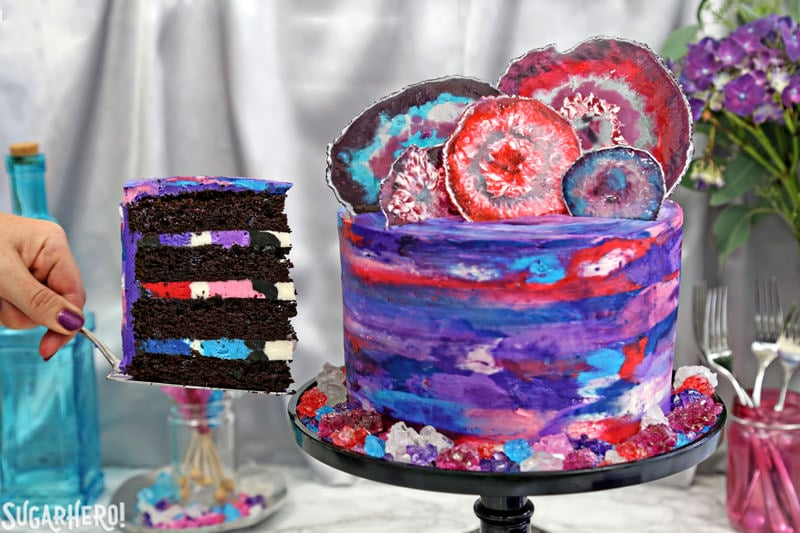 Removing a slice of chocolate cake from the side of a fully decorated agate cake, with a watercolor buttercream effect and candy agate slices on top | From SugarHero.com