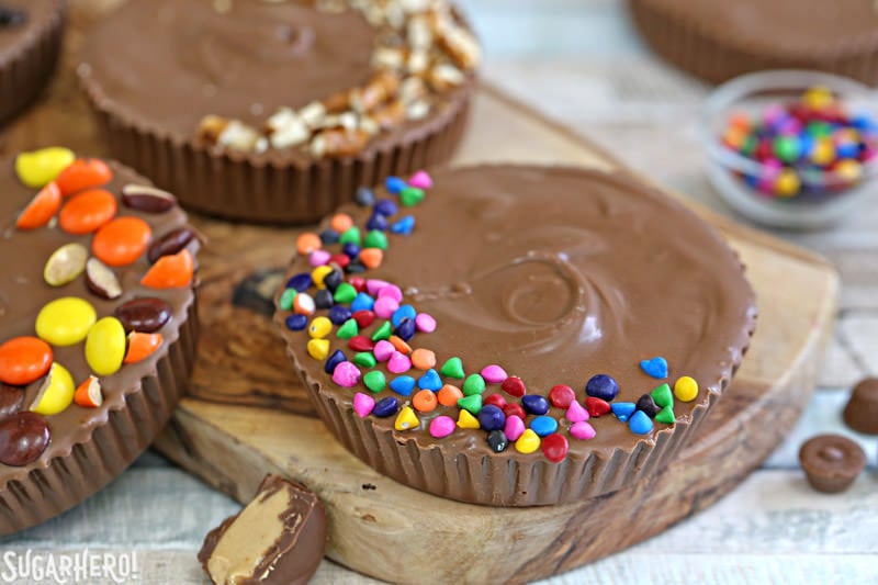 Mega Stuffed Peanut Butter Cups - jumbo homemade peanut butter cups, filled with goodies like candy, cookies, pretzels, or potato chips! | From SugarHero.com Mega Stuffed Peanut Butter Cups - giant peanut butter cup on a wooden tray with colorful rainbow chips on top | From SugarHero.com