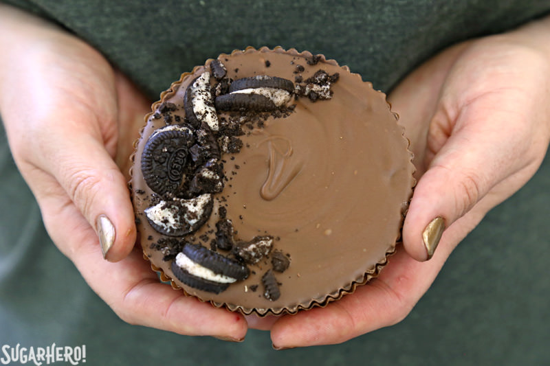 Mega Stuffed Peanut Butter Cups - hands holding a giant peanut butter cup topped with crushed Oreos | From SugarHero.com