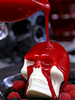 Raspberry ganache being poured over white chocolate skull on a black plate with raspberries.