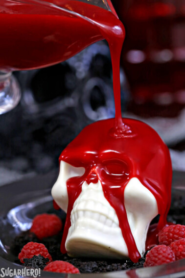 Raspberry ganache being poured over white chocolate skull on a black plate with raspberries.