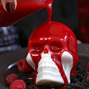 Raspberry ganache being poured over white chocolate skull on a black plate with raspberries.