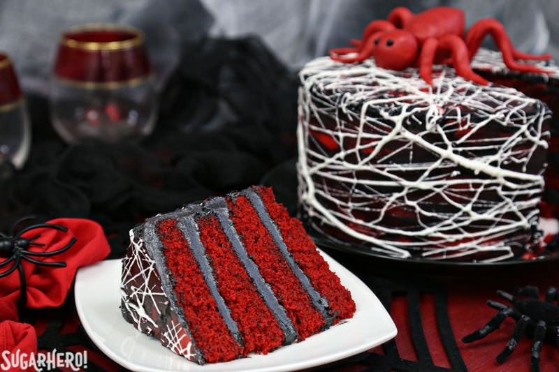 Red Velvet Marshmallow Spiderweb Cake—classic red velvet cake with black chocolate buttercream, covered with a web of marshmallow spiderwebs! | From SugarHero.com Red Velvet Marshmallow Spiderweb Cake - slice of cake on a plate with full cake behind it | From SugarHero.com
