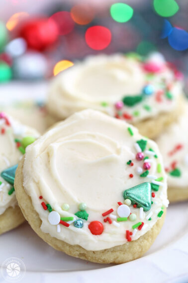 Five big soft sugar cookies with red, white, and green sprinkles and Christmas lights in the background.