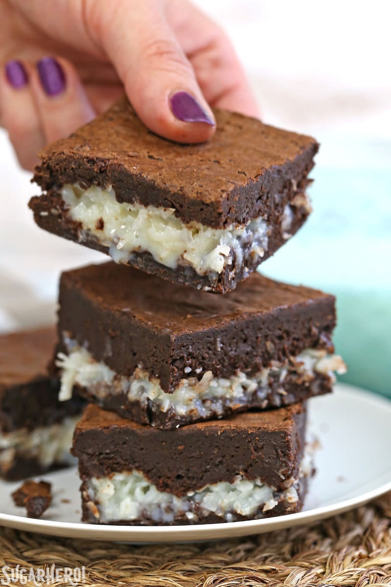 The BEST Coconut Brownies - hand picking up a gooey coconut brownie from a stack of brownies | From SugarHero.com