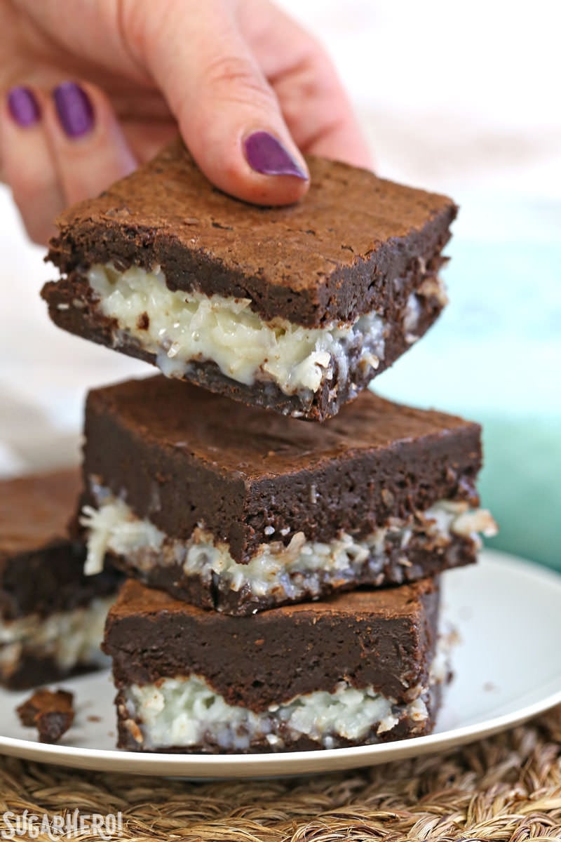 These are the BEST Coconut Brownies! Rich, fudgy brownies and a gooey coconut center. Better than a candy bar! | From SugarHero.com The BEST Coconut Brownies - hand picking up a gooey coconut brownie from a stack of brownies | From SugarHero.com