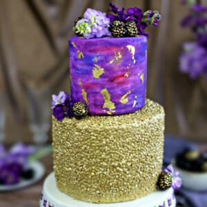 Close up of a Gold Sequin Watercolor Cake with flowers in the background.