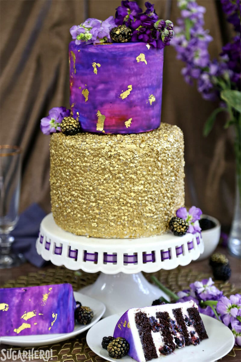 Gold Sequin Watercolor Cake - a stunning layer cake decorated with completely edible gold sequins and a gorgeous watercolor fondant effect! Perfect for parties, showers, or as a nontraditional wedding cake! | From SugarHero.com Gold Sequin Watercolor Cake - picture of the full cake on a cake stand with several slices scattered around it | From SugarHero.com