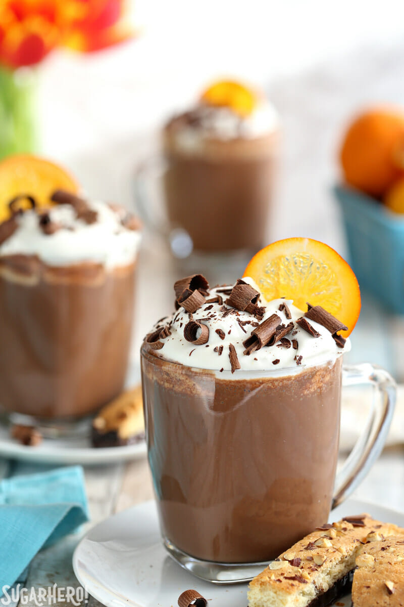 Orange Hot Chocolate mugs topped with whipped cream, chocolate curls, and a candied orange slice | From SugarHero.com