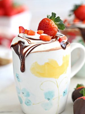 Strawberry hot chocolate in a floral mug with whipped cream and sliced strawberries on top next to chocolate covered strawberries.