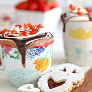 Strawberry hot chocolate in a floral mug with whipped cream and sliced strawberries on top next to jam cookies and a cup in the background.