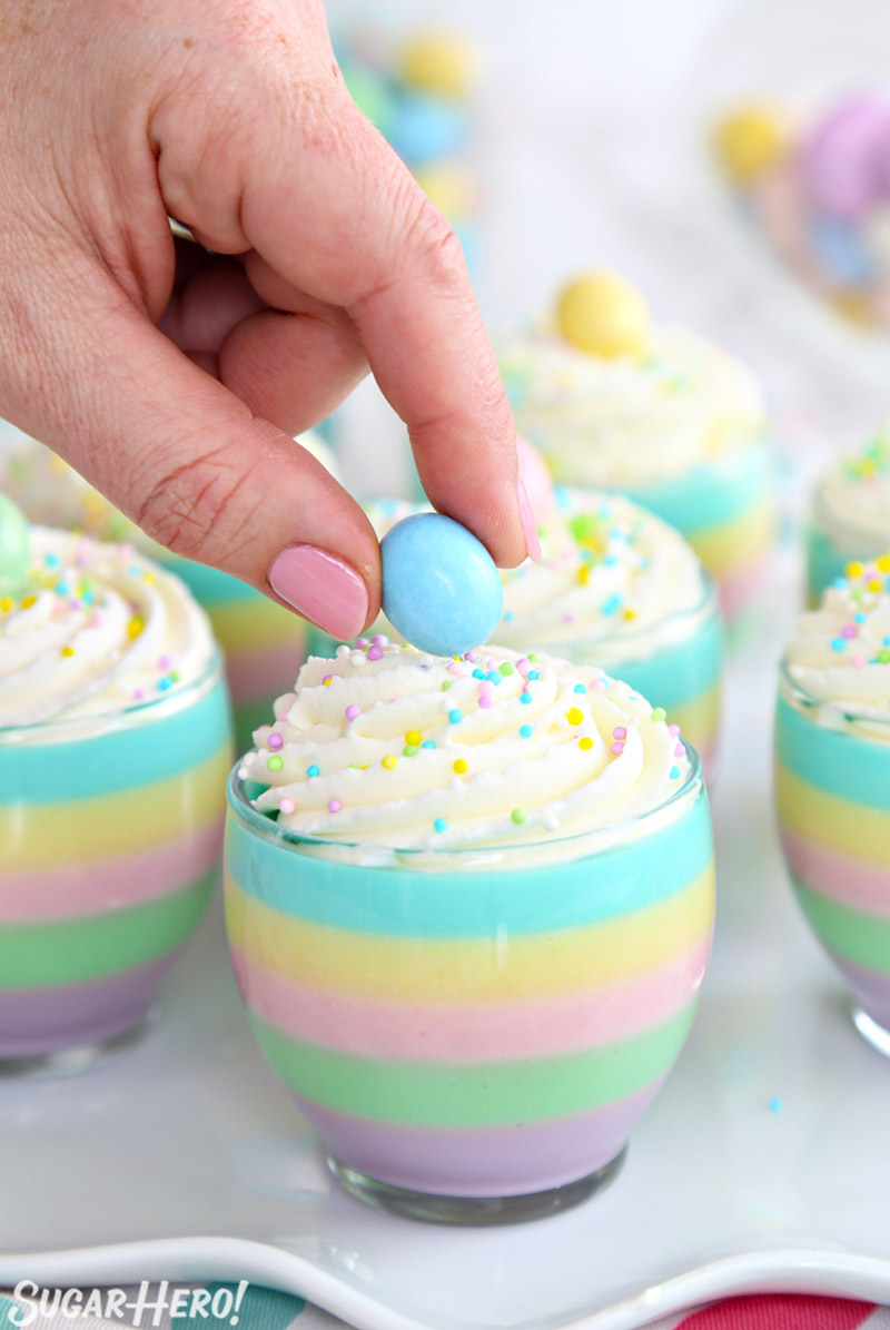 A hand placing a blue Easter egg decoration on top of a striped pastel gelatin cup.