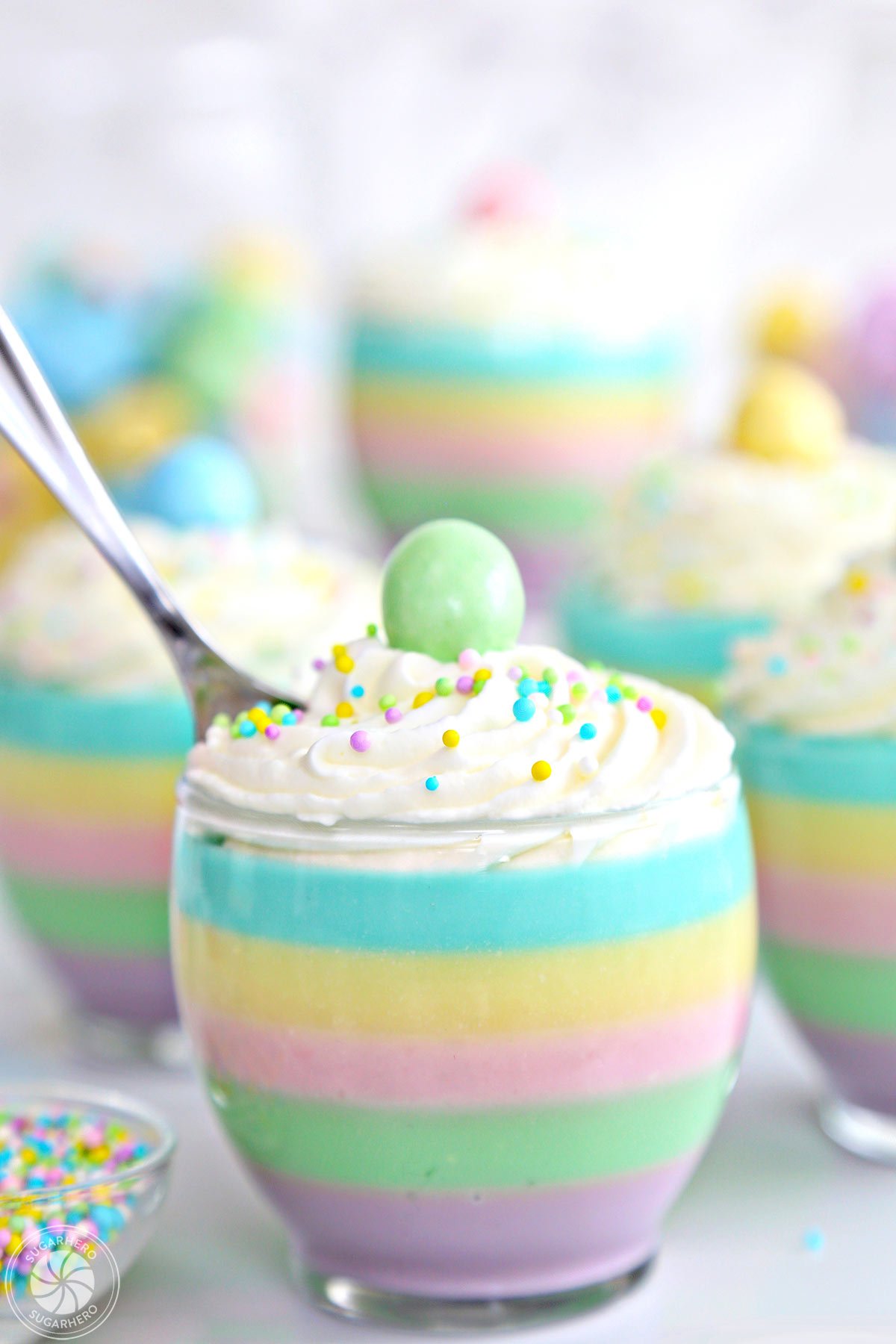 Pastel Rainbow Gelatin Cups – an easy spring dessert that’s simple, kid-friendly, and so beautiful! Serve them plain, or top them with whipped cream and sprinkles! | From SugarHero.com Close-up of striped pastel gelatin cup with a spoon sticking out of the top