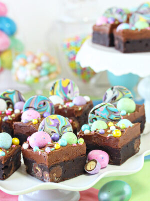 Eight Easter Egg Brownies topped with chocolate frosting and pastel chocolate eggs and sprinkles on a white scalloped cake stand and another cake stand in the background with a few more Easter Egg Brownies decorated in a similar fashion.