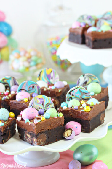 Frosted Easter Brownies topped with chocolate frosting and pastel chocolate eggs and sprinkles on a white scalloped cake stand and another cake stand in the background with a few more Easter Egg Brownies decorated in a similar fashion.