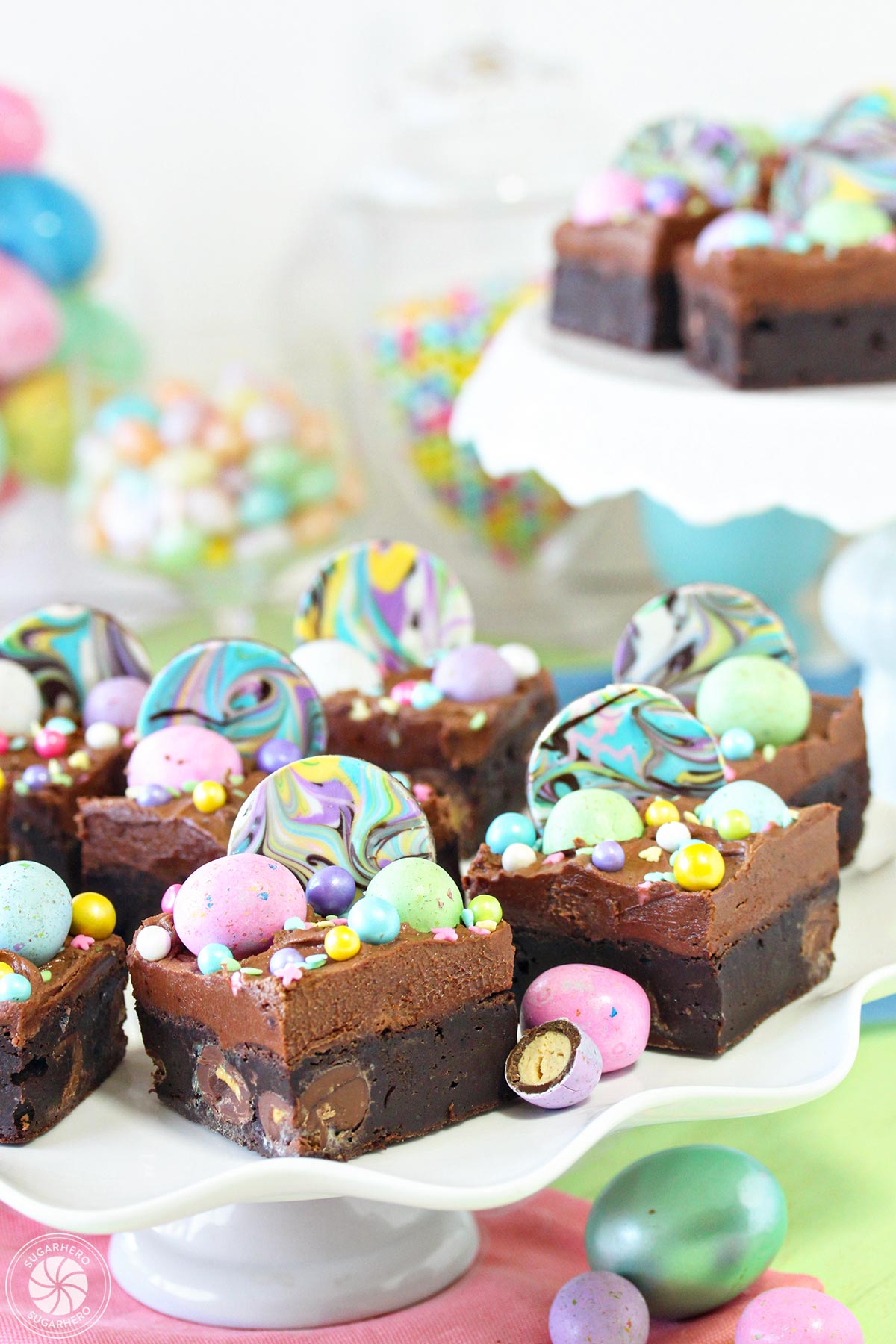 Eight Easter Egg Brownies topped with chocolate frosting and pastel chocolate eggs and sprinkles on a white scalloped cake stand and another cake stand in the background with a few more Easter Egg Brownies decorated in a similar fashion.