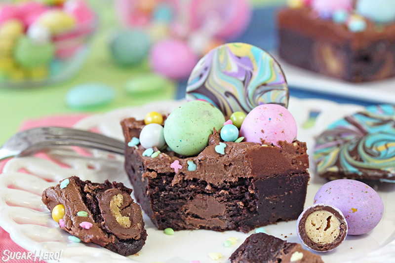 An Easter Egg Brownie covered in chocolate frosting and topped with pastel chocolate eggs, assorted sprinkles, and swirled chocolate bark with a bite missing and set beside it.