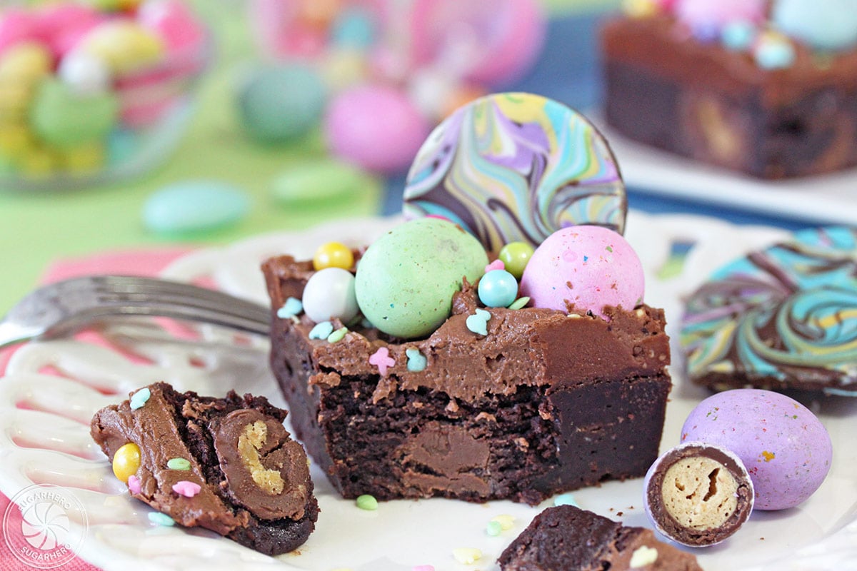 An Easter Egg Brownie covered in chocolate frosting and topped with pastel chocolate eggs, assorted sprinkles, and swirled chocolate bark with a bite missing and set beside it.