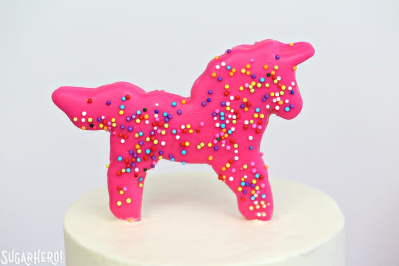 Circus Animal Layer Cake - a big, bright pink cake with funfetti cake layers, chopped up circus animal cookies inside, and a cool upside-down ganache drip decoration! | From SugarHero.com Circus Animal Layer Cake - close-up of pink sprinkled unicorn cookie on top of circus animal cake | From SugarHero.com