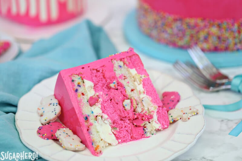 Circus Animal Layer Cake - a big, bright pink cake with funfetti cake layers, chopped up circus animal cookies inside, and a cool upside-down ganache drip decoration! | From SugarHero.com Circus Animal Layer Cake - slice of pink and white cake on a plate | From SugarHero.com