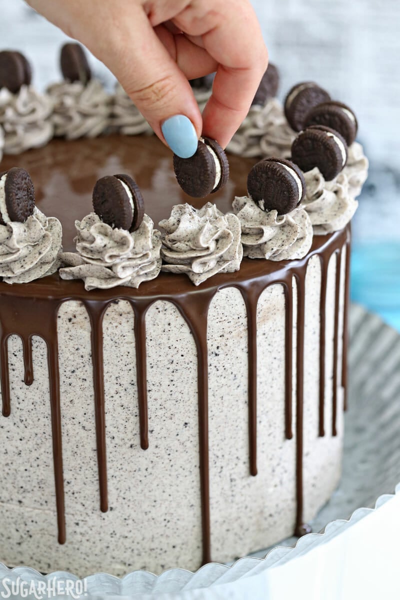 Cookies and Cream Cake - this epic cookies and cream layer cake will satisfy ALL of your chocolate cravings! It has rich devil's food cake plus buttercream with lots of cookie crumbs mixed right in. | From SugarHero.com Cookies and Cream Cake - hand placing mini Oreo on top of cake | From SugarHero.com