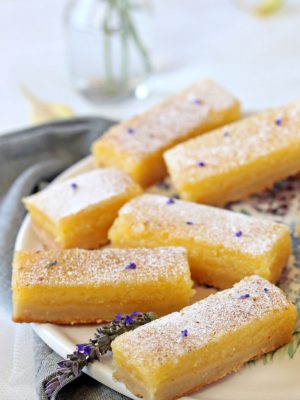 Six lavender lemon bars on a round white plate with a lavender flower on it.