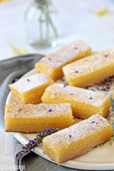 Six lavender lemon bars on a round white plate with a lavender flower on it.