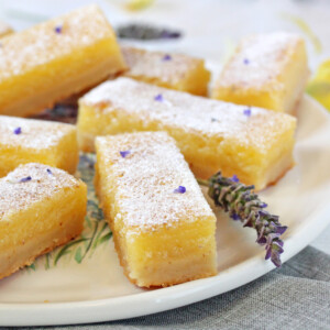 Seven lavender lemon bars on a round white plate with a lavender flower next to them.