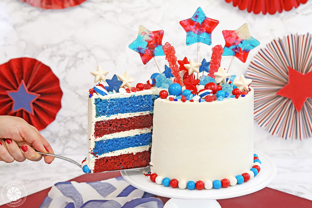 Fourth of July Layer Cake - celebrate Independence Day with a bang by making this fun July 4th cake! Red and blue velvet cake, smooth buttercream, and tons of fun candies on top--what more do you need for a party?! | From SugarHero.com Fourth of July Layer Cake - hand removing a slice of red and blue velvet cake with a cake server | From SugarHero.com